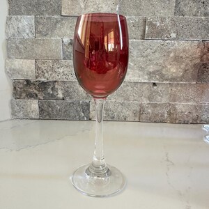 May include: A single wine glass with a ruby-colored bowl and a clear, slender stem. The glass sits on a clear, circular base. The background features a light-colored countertop and a gray stone tile wall.