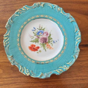 May include: A decorative plate with a turquoise rim and a white center. The center features a floral arrangement with red, blue, and purple flowers. Gold accents adorn the rim and the floral design, creating an elegant aesthetic.