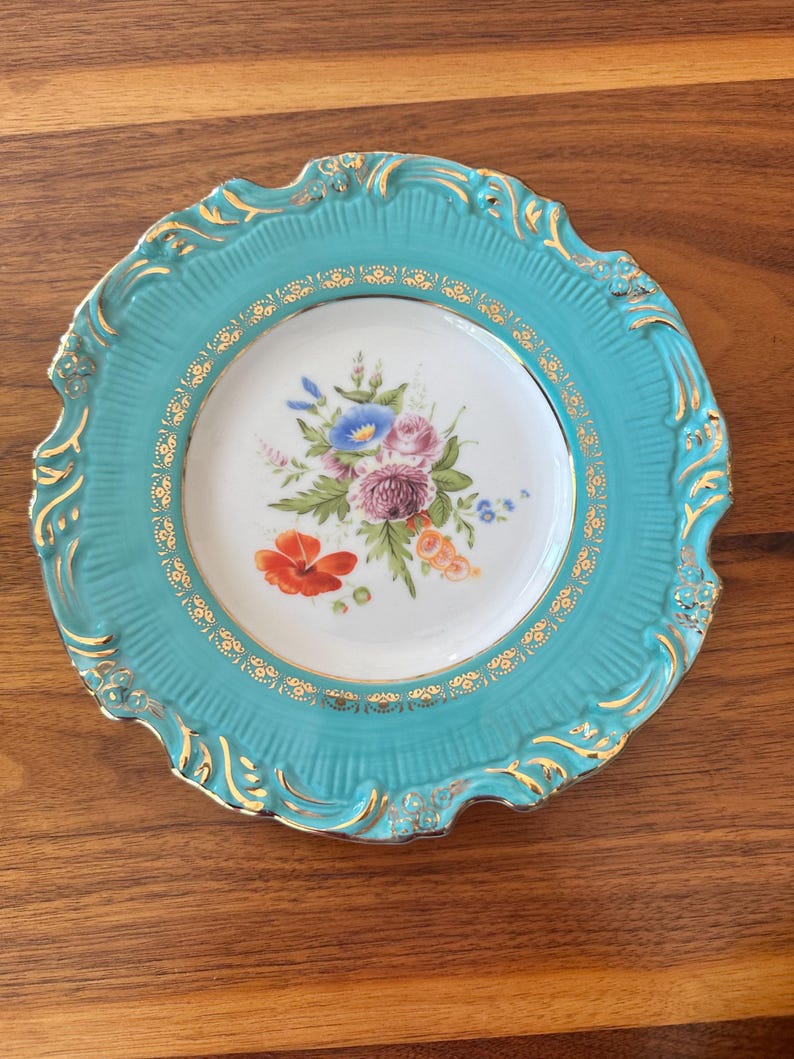 May include: A decorative plate with a turquoise rim and a white center. The center features a floral arrangement with red, blue, and purple flowers. Gold accents adorn the rim and the floral design, creating an elegant aesthetic.