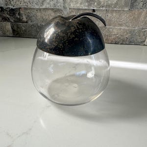 Puede incluir: Un tarro de vidrio transparente con una tapa de manzana plateada. La tapa tiene una superficie texturizada y un pequeño tallo.