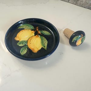 Może przedstawiać: Ciemnoniebieski ceramiczny zestaw talerza i korka do butelki. Talerz i korek są ozdobione żółtymi cytrynami i zielonymi liśćmi. Korek ma korkową podstawę.