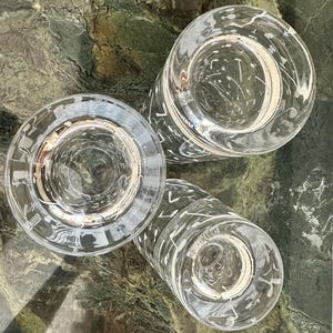 May include: Three clear glass tumblers arranged in a cluster, each featuring a white printed pattern of various musical notes and symbols scattered across the exterior surface, viewed from a top-down perspective showing the circular rims.