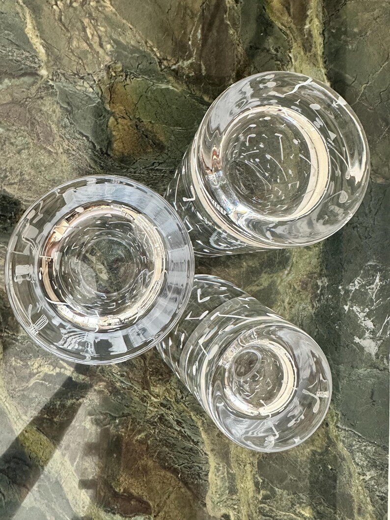 May include: Three clear glass tumblers arranged in a cluster, each featuring a white printed pattern of various musical notes and symbols scattered across the exterior surface, viewed from a top-down perspective showing the circular rims.