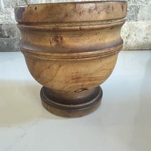 Könnte beinhalten: Ein hellbrauner Holz-Mörser und Stößel. Die Schüssel ist abgerundet und steht auf einem Sockel. Die Holzmaserung ist sichtbar, und der Mörser hat eine glatte, polierte Oberfläche. Er ist zum Mahlen und Mischen von Zutaten bestimmt.