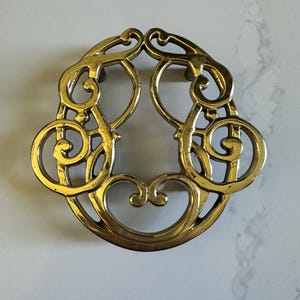 May include: Ornate, gold-colored metal trivet with a symmetrical design. The trivet features a series of swirling, scroll-like patterns and heart shapes. The metal has a slightly aged appearance, suggesting it is vintage or antique.