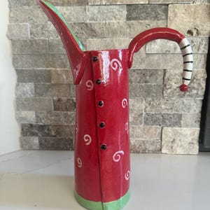 May include: A red ceramic pitcher with a long spout and a curved handle. The pitcher is decorated with white swirls and black buttons down the front. The handle has black and white stripes. The base and the inside of the spout are green.