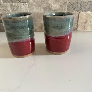 Puede incluir: Dos vasos de cerámica hechos a mano. La mitad superior de cada vaso está esmaltada en color verde azulado, pasando a un rojo vibrante en la parte inferior. La base de cada vaso es de color arcilla natural sin esmaltar.
