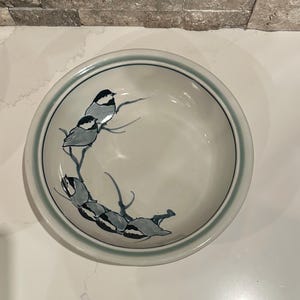 Puede incluir: Un cuenco de cerámica con un borde azul claro y un diseño pintado a mano de pájaros posados en ramas. Las aves están representadas en tonos grises y negros, añadiendo un toque de arte inspirado en la naturaleza al diseño del cuenco.