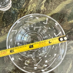 May include: A clear glass tumbler featuring a white printed pattern of golf clubs and golf balls. A yellow measuring tape is placed across the top opening, indicating a diameter of approximately 3.25 inches for the rim of the glass.