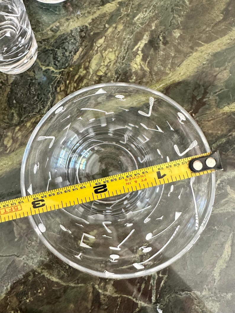 May include: A clear glass tumbler featuring a white printed pattern of golf clubs and golf balls. A yellow measuring tape is placed across the top opening, indicating a diameter of approximately 3.25 inches for the rim of the glass.