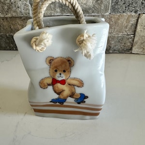 Può includere: Un contenitore in ceramica bianca a forma di borsa con due spessi manici in corda beige annodati sul davanti. La parte anteriore presenta un orsacchiotto marrone con papillon rosso e pattini a rotelle blu, con due strisce marroni alla base.