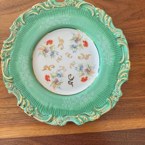 May include: A decorative plate with a mint green rim and a white center. The rim has a gold filigree design. The center features a floral pattern with red, purple, and blue flowers. A unique piece for display.