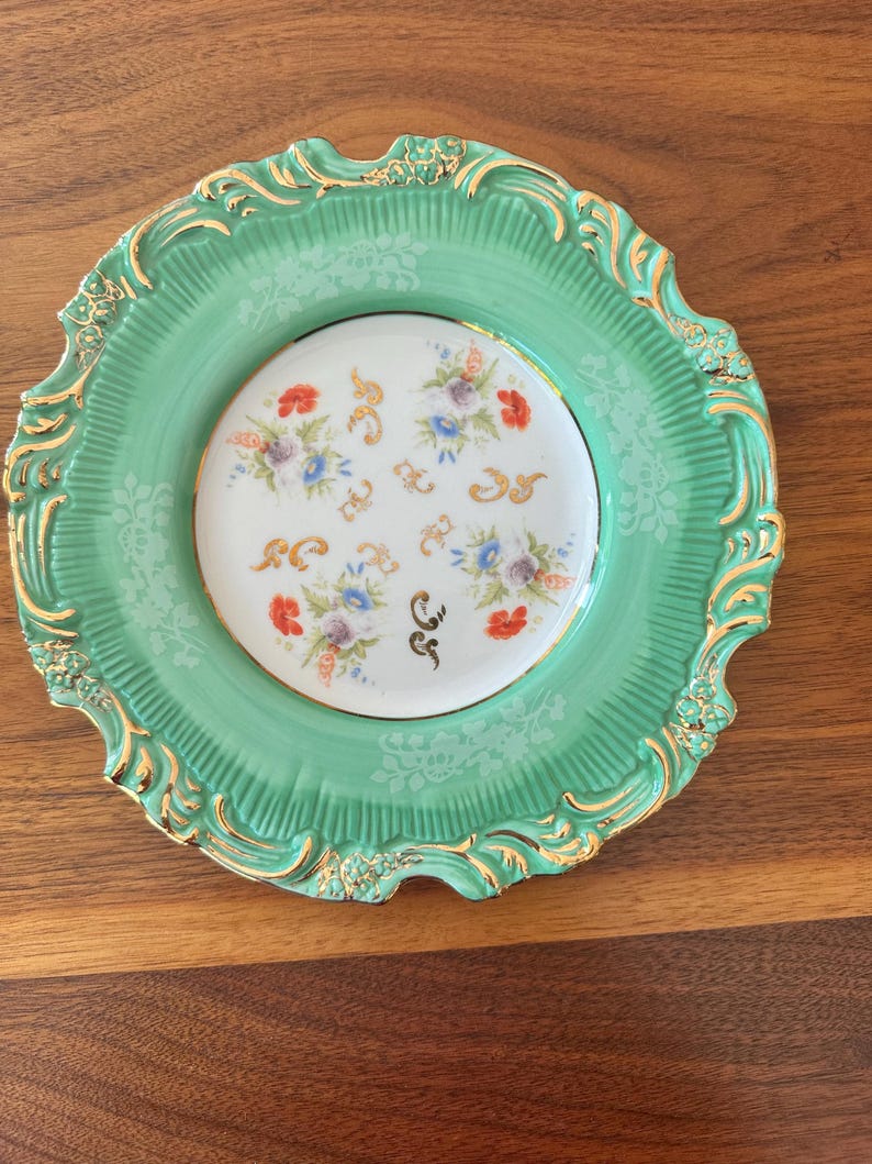 May include: A decorative plate with a mint green rim and a white center. The rim has a gold filigree design. The center features a floral pattern with red, purple, and blue flowers. A unique piece for display.