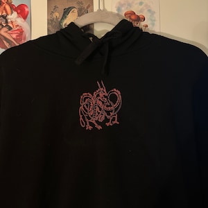 Könnte beinhalten: Schwarzer Kapuzenpullover mit Kordelzug und einem roten, gestickten Design auf der Vorderseite. Das Design scheint eine stilisierte Kreatur zu sein. Der Kapuzenpullover hängt an einem weißen Kleiderbügel vor einer weißen Wand mit Kunstwerken.