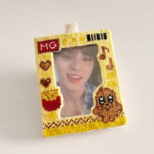 May include: A handmade yellow photo frame with a picture of a person. The frame is decorated with embroidered details, including a heart, french fries, a musical note, and a dog. The letters "MG" are embroidered on the top left.