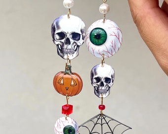 May include: A pair of Halloween-themed dangle earrings with a skull, pumpkin, eyeball, and spiderweb design. The earrings are gold-toned and feature white pearl accents.