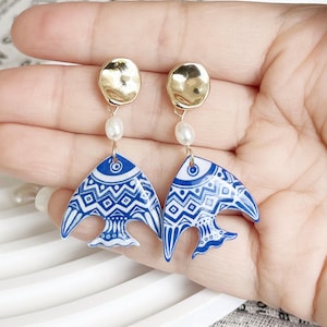 Pode incluir: Um par de brincos com uma parte superior dourada com textura martelada, uma pequena pérola branca e um pingente em forma de peixe azul e branco. O pingente de peixe tem um design geométrico.