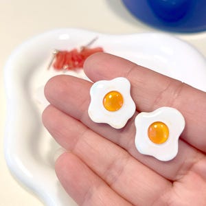 Peut inclure: Une paire de boucles d'oreilles en forme d'œuf au plat, blanches et jaunes. Les boucles d'oreilles sont en forme d'œuf au plat avec un jaune blanc et un jaune jaune.