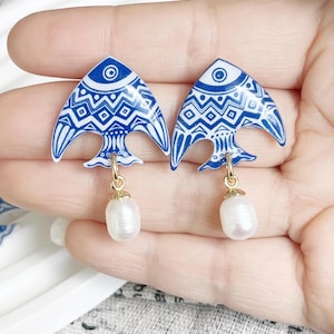 Pode incluir: Um par de brincos azuis e brancos em forma de peixe com padrões intrincados. Cada brinco tem um gancho dourado e uma pérola branca pendurada. Os brincos são segurados por uma mão.