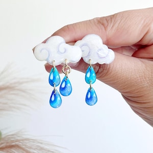 May include: Earrings shaped like clouds with blue teardrops. The earrings have white clouds with light purple detailing and gold-coloured hooks. The blue teardrops have a gradient effect, going from a darker to a lighter shade of blue.