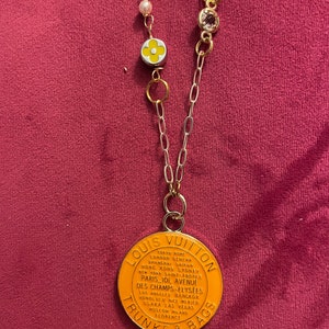May include: A gold chain necklace with a round orange pendant that says "Louis Vuitton Trunks & Bags" with a list of cities around the edge. The pendant has a white flower charm attached to it.