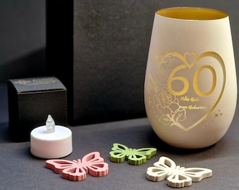May include: A white candle holder with a gold rim and a cutout design of a heart with the number 60 inside. The text "Alles Gute zum Geburtstag" is written inside the heart. The candle holder is sitting on a black surface with a small white tea light and three wooden butterflies.