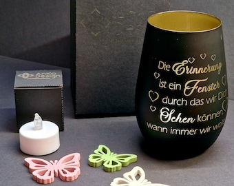 May include: Black glass candle holder with a gold rim and the text "Die Erinnerung ist ein Fenster durch das wir Dich sehen können wann immer wir wollen" in gold lettering. The candle holder is surrounded by three small wooden butterflies in pink, green, and white.
