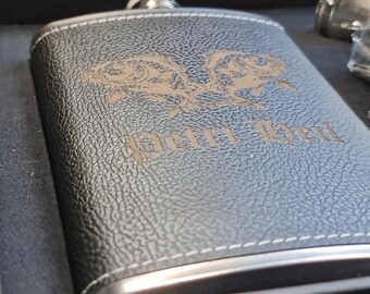 May include: A black leather-covered flask with a silver rim and a design of two fish with the text "Happy Hour" engraved on it. The flask is in a black box.