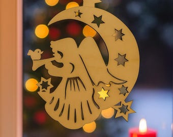May include: A wooden Christmas ornament featuring an angel playing a horn, a crescent moon, and stars. The ornament hangs in front of a window with bokeh lights and three red candles.
