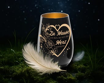 May include: A black stemless wine glass with a gold rim and a heart-shaped design. The design includes roses and the German text "Zu Hause ist dort, wo sich das Herz wohlfühlt." A white feather rests near the glass.