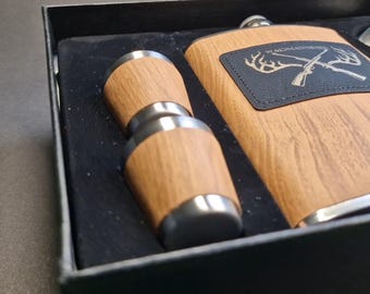 May include: A wooden gift box with a stainless steel flask and two shot glasses. The flask has a black leather patch with the text "WADMAN" and a deer antler design.