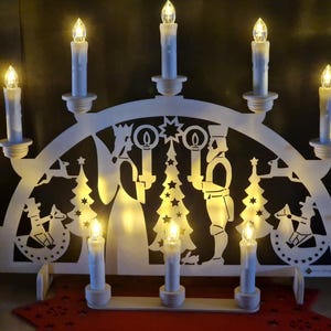 Könnte beinhalten: Eine weiße Weihnachts-Kerzenbrücke aus Holz mit neun LED-Kerzen. Die Brücke zeigt ausgeschnittene Silhouetten von Engeln, Weihnachtsbäumen und anderen festlichen Figuren. Die Kerzen sind in einer Bogenform angeordnet und erzeugen ein warmes, einladendes Leuchten.