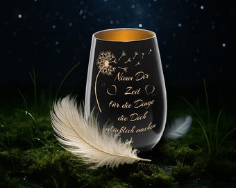 May include: A black stemless wine glass with a gold rim and a dandelion design. The glass has the text "Nimm Dir Zeit für die Dinge die Dich glücklich machen". A white feather rests on the mossy surface.