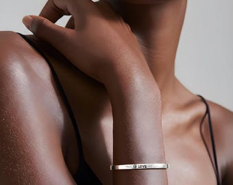 May include: A silver-colored cuff bracelet with the word "LOVE" engraved on it. The bracelet is worn on a person's wrist. The person is wearing a black top.