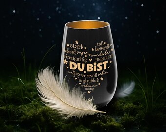 May include: A black stemless wine glass with a gold interior rim. The glass is decorated with gold text and star accents. A white feather rests on the mossy ground in front of the glass.