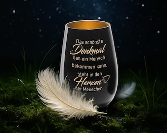 May include: A black stemless wine glass with gold trim, featuring the text "Das schönste Denkmal..." in elegant script. A white feather rests on the mossy ground. The glass is a memorial or remembrance item.