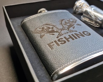 May include: Black leather flask with a silver cap, featuring an engraved design of two fish and the word "FISHING". The flask is presented in a black gift box, with a silver accessory visible in the background.