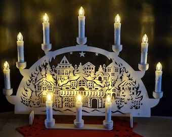 May include: A white wooden Christmas candle bridge with a cut-out village scene. The bridge holds ten electric candles with warm yellow lights. The base is on a red surface.