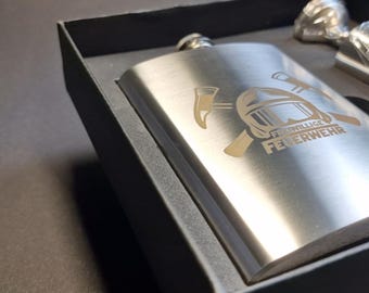 May include: A silver flask with a black box. The flask has an engraved design of crossed axes and a firefighter's helmet with the text "Freiwillige Feuerwehr".