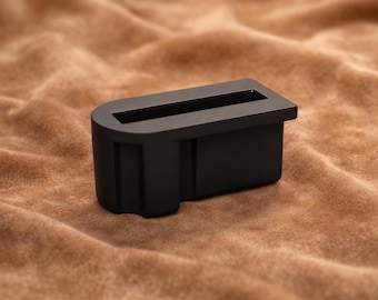 May include: A matte black rectangular object with a recessed slot, resting on a brown, textured surface. The object has a rounded base and a rectangular opening. The overall shape is a stylized U.
