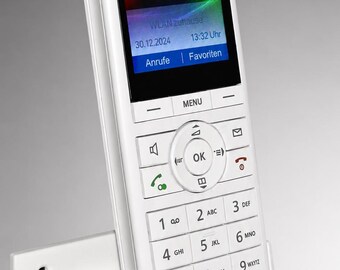May include: A white cordless DECT telephone with a color screen displaying the text FRITZ!Fon, WLAN zuhause, 30.12.2024, 13:32 Uhr, Anrufe, and Favoriten. The device features a standard numeric keypad, navigation buttons, and a matching white charging base.
