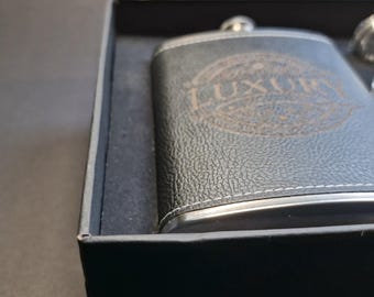May include: A black leather-covered flask with a silver rim and a silver cap. The flask has the word "Luxury" engraved on the front.