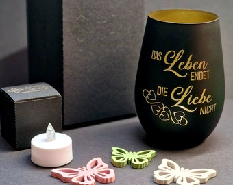 May include: A black glass candle holder with gold lettering that reads "Das Leben endet, die Liebe nicht". The candle holder is surrounded by three small butterfly decorations in pink, green, and white.