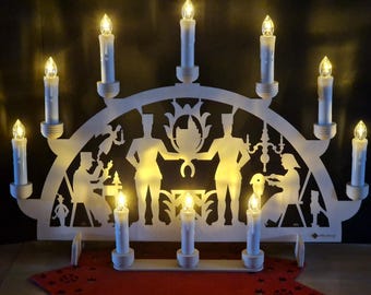 May include: White wooden candle bridge with nine LED candles. The bridge features cut-out silhouettes of figures and decorative elements. The candles are lit, creating a warm, inviting glow. A red decorative mat is at the base.