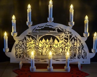 May include: A white wooden Christmas candle bridge with nine electric candles. The bridge features a cut-out design of a village scene with houses, trees, and a train. The candles are lit, creating a warm, inviting glow.