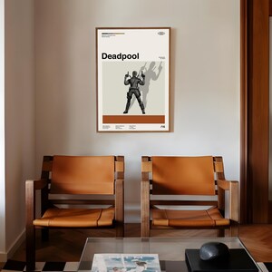 May include: A framed poster featuring the word "Deadpool" in bold black letters. The poster depicts a silhouetted figure holding two guns, with a shadow cast behind. The poster has a cream background with brown and black accents.