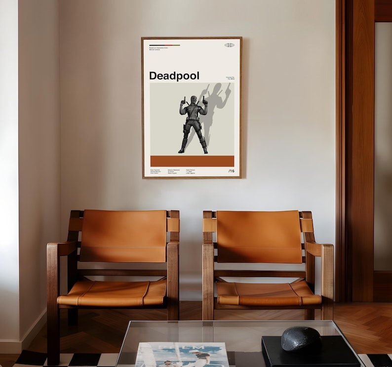 May include: A framed poster featuring the word "Deadpool" in bold black letters. The poster depicts a silhouetted figure holding two guns, with a shadow cast behind. The poster has a cream background with brown and black accents.