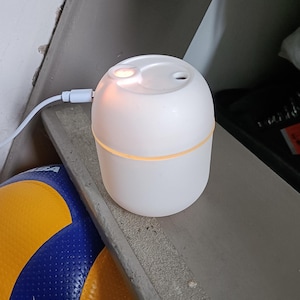 Puede incluir: Un difusor eléctrico blanco, redondo, con una luz encendida, sentado en un estante con un balón de voleibol amarillo y azul en primer plano.