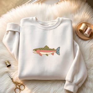 May include: A white crewneck sweatshirt featuring a central embroidered graphic of a trout fish. The fish has a green back, a pink lateral stripe, and a blue tail fin decorated with small white stars. The sweatshirt has long sleeves with cuffs.