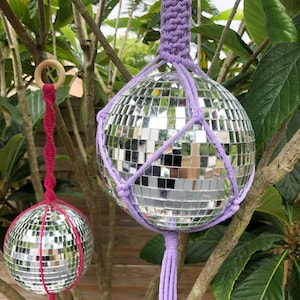 May include: Two disco ball hanging planters. One is suspended by a purple macrame hanger, the other by a pink macrame hanger. The disco balls are round and covered in small mirrored squares. The planters hang from tree branches.